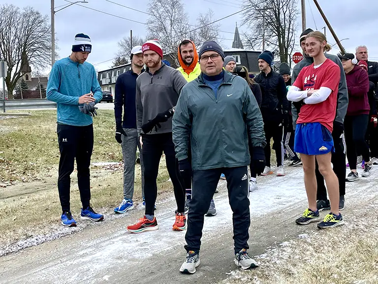 Pete Rettler stands in the foreground, dressed in winter running gear, ready to participate in the 2023 Rettler Run. He is surrounded by other runners dressed in cold-weather clothing, some with hats and gloves, all preparing to begin the run on a snowy path. The overcast sky and bare trees in the background suggest a chilly day